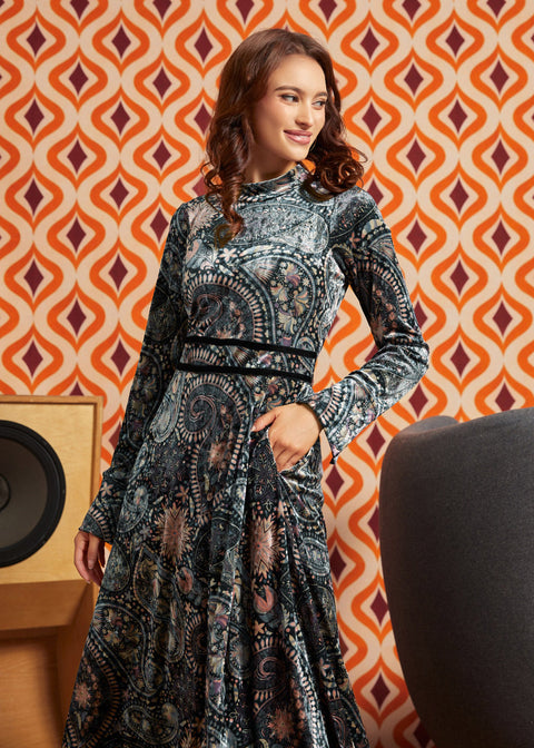 Woman wearing dark paisley floral midi dress with long flared sleeves and black waist trim, front view with one hand in side pocket