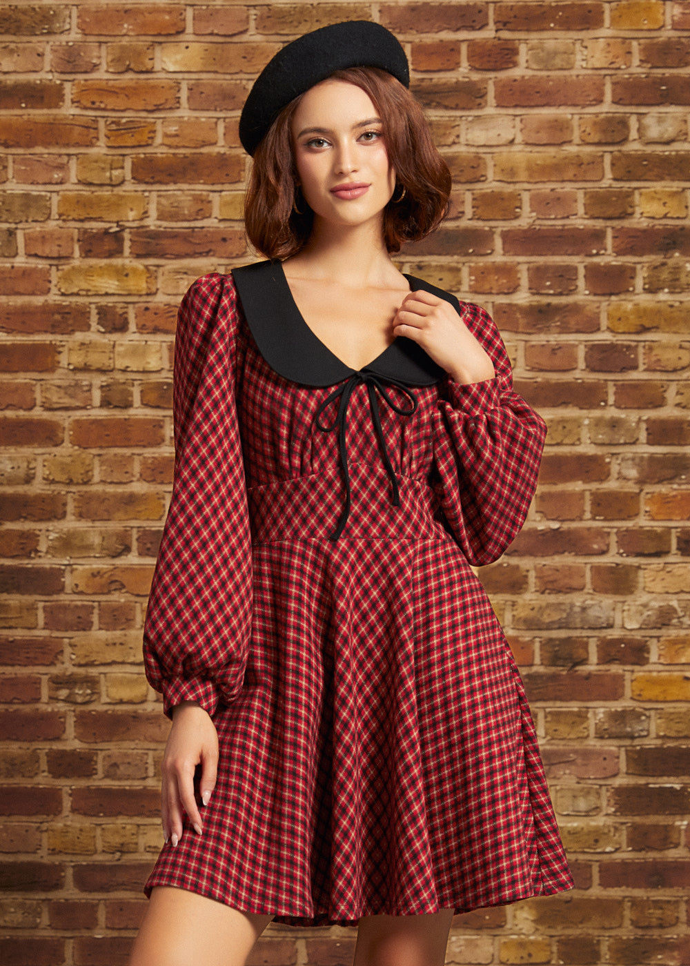 A woman wearing a red and black checkered long-sleeve dress with a black Peter Pan collar and a black beret, posing against a rustic brick wall.