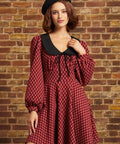 A woman wearing a red and black checkered long-sleeve dress with a black Peter Pan collar and a black beret, posing against a rustic brick wall.