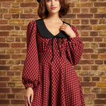 A woman wearing a red and black checkered long-sleeve dress with a black Peter Pan collar and a black beret, posing against a rustic brick wall.