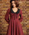 A woman wearing a red and black checkered long-sleeve dress with a black Peter Pan collar and a black beret, standing against a brick wall.