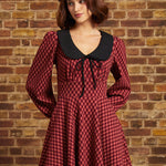 A woman wearing a red and black checkered long-sleeve dress with a black Peter Pan collar and a black beret, standing against a brick wall.