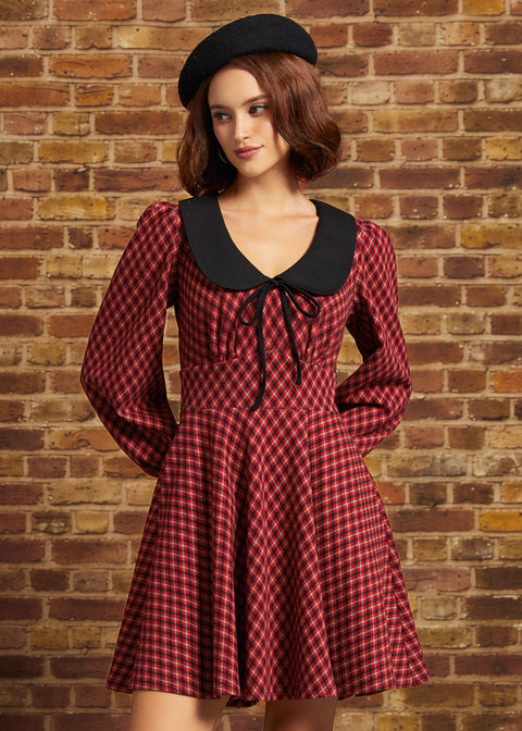 A woman wearing a red and black checkered long-sleeve dress with a black Peter Pan collar and a black beret, standing against a brick wall.