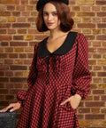 A woman wearing a red and black checkered long-sleeve dress with a large black Peter Pan collar and a black beret, standing against a brick wall.