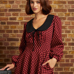 A woman wearing a red and black checkered long-sleeve dress with a large black Peter Pan collar and a black beret, standing against a brick wall.