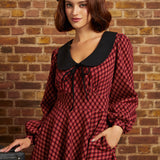 A woman wearing a red and black checkered long-sleeve dress with a large black Peter Pan collar and a black beret, standing against a brick wall.