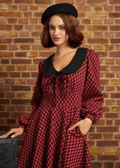 A woman wearing a red and black checkered long-sleeve dress with a large black Peter Pan collar and a black beret, standing against a brick wall.