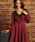 A woman wearing a red and black checkered long-sleeve dress with a black Peter Pan collar and black beret, posing in front of a brick wall next to a vintage suitcase.
