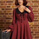 A woman wearing a red and black checkered long-sleeve dress with a black Peter Pan collar and black beret, posing in front of a brick wall next to a vintage suitcase.