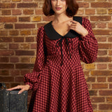 A woman wearing a red and black checkered long-sleeve dress with a black Peter Pan collar and black beret, posing in front of a brick wall next to a vintage suitcase.