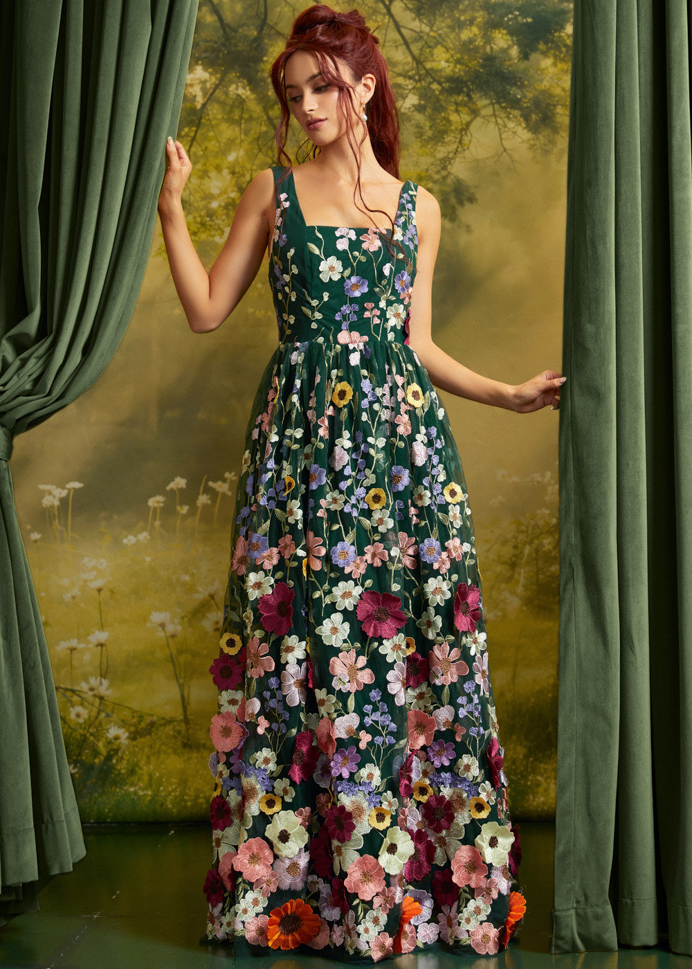A woman stands between green curtains wearing a sleeveless dark green maxi dress covered in large multicolored floral appliqués. The background is a painterly yellow-green landscape scene.