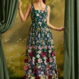 A woman stands between green curtains wearing a sleeveless dark green maxi dress covered in large multicolored floral appliqués. The background is a painterly yellow-green landscape scene.