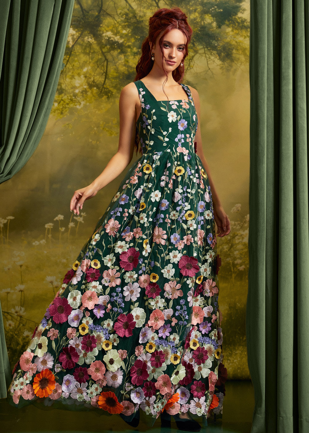 A woman wearing a sleeveless dark green maxi dress covered in colorful floral embroidery, posed in front of a scenic backdrop with green curtains.