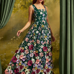 A woman wearing a sleeveless dark green maxi dress covered in colorful floral embroidery, posed in front of a scenic backdrop with green curtains.