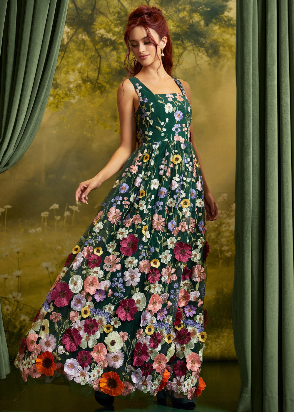 A woman wearing a sleeveless, floor-length floral dress with a dark green base and large multicolored flowers, posed in front of a scenic backdrop with green curtains.