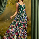 A woman wearing a sleeveless, floor-length floral dress with a dark green base and large multicolored flowers, posed in front of a scenic backdrop with green curtains.