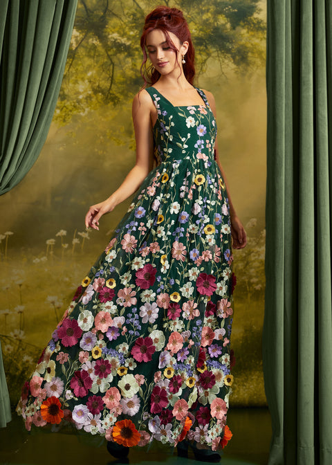 A woman wearing a sleeveless, floor-length floral dress with a dark green base and large multicolored flowers, posed in front of a scenic backdrop with green curtains.