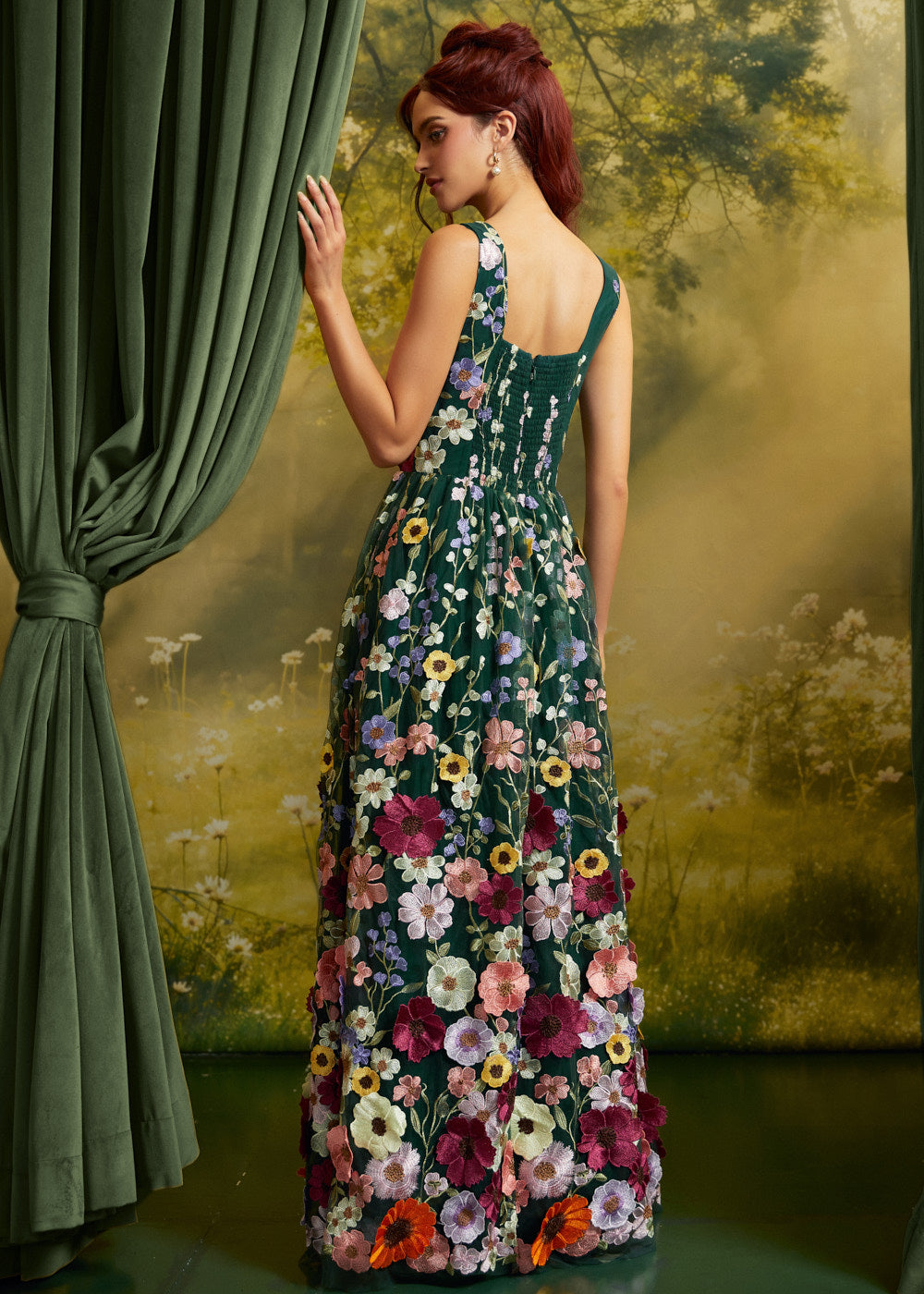 A woman stands with her back to the camera wearing a sleeveless dark green maxi dress covered in colorful floral embroidery, posed in front of a green curtain and a scenic forest backdrop.