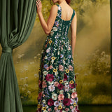 A woman stands with her back to the camera wearing a sleeveless dark green maxi dress covered in colorful floral embroidery, posed in front of a green curtain and a scenic forest backdrop.