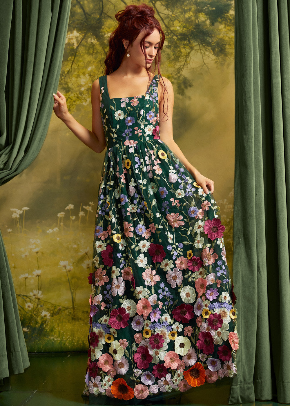 A woman wearing a sleeveless, floor-length floral dress with a dark green base and large multicolored flowers, posed in front of a scenic backdrop with green curtains.