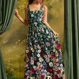 A woman wearing a sleeveless, floor-length floral dress with a dark green base and large multicolored flowers, posed in front of a scenic backdrop with green curtains.