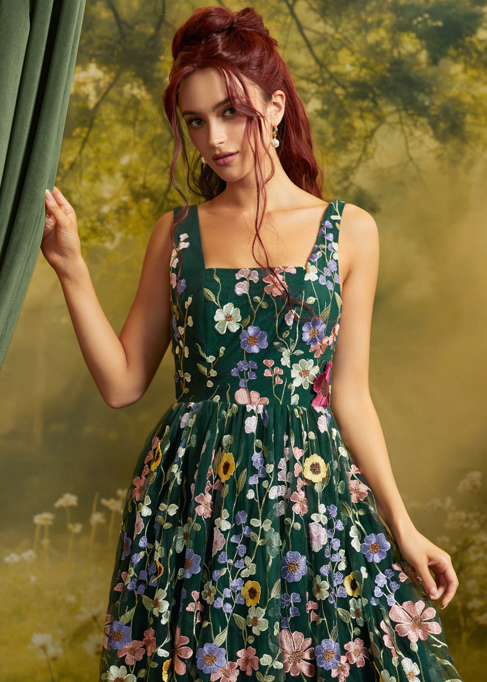 A woman with red hair wearing a sleeveless dark green ModCloth dress with colorful floral embroidery, posed in front of a soft-focus green and yellow backdrop.