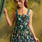 A woman with red hair wearing a sleeveless dark green ModCloth dress with colorful floral embroidery, posed in front of a soft-focus green and yellow backdrop.