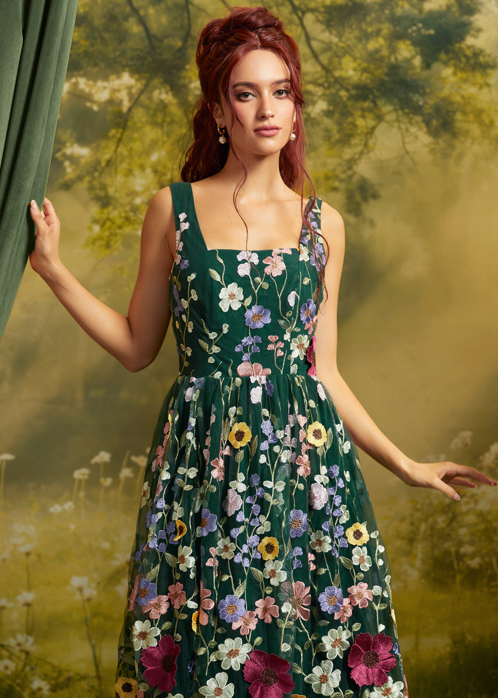 A woman modeling a sleeveless dark green formal dress with colorful floral embroidery, posed against a soft-focus yellow-green outdoor background with a green curtain on the left.