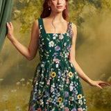 A woman modeling a sleeveless dark green formal dress with colorful floral embroidery, posed against a soft-focus yellow-green outdoor background with a green curtain on the left.