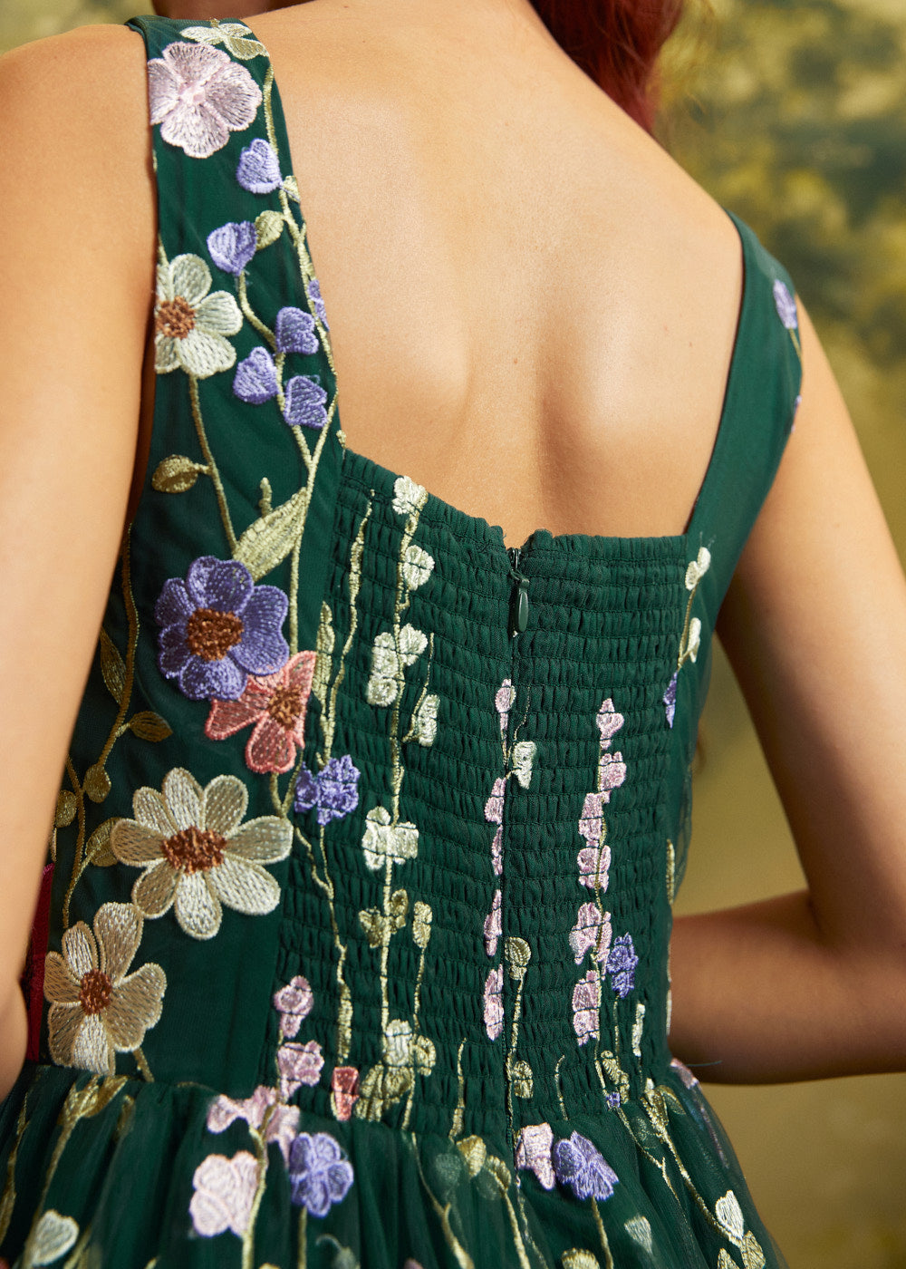 Close-up back view of a dark green sleeveless dress with floral embroidery and a smocked bodice, shown on a model.