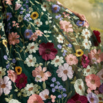 A close-up of a flowing dark green embroidered dress with large multicolored floral motifs in pink, burgundy, purple, yellow, and white, worn by a model whose hand and arm are visible.