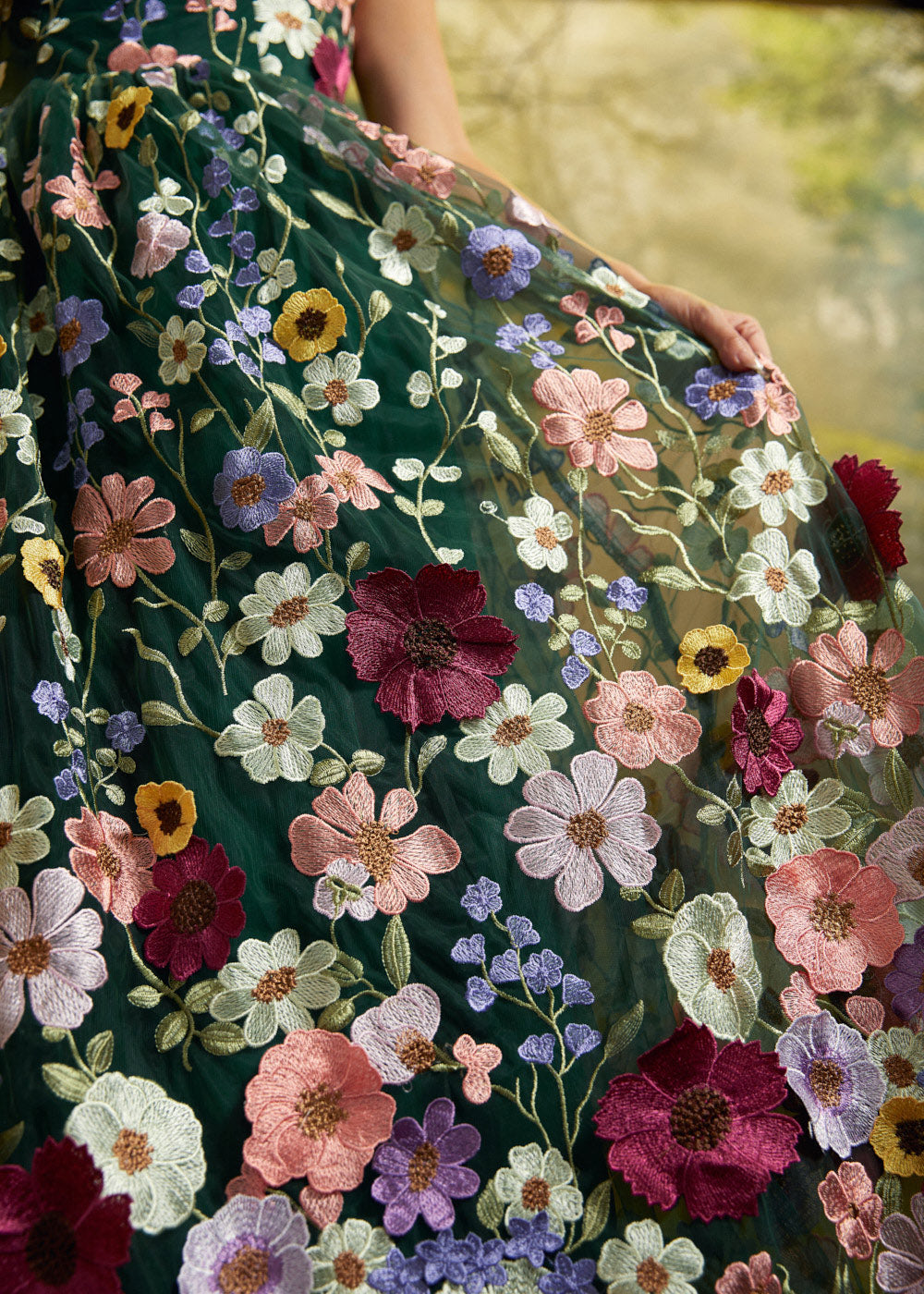 A close-up of a flowing dark green embroidered dress with large multicolored floral motifs in pink, burgundy, purple, yellow, and white, worn by a model whose hand and arm are visible.