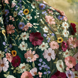 A close-up of a flowing dark green embroidered dress with large multicolored floral motifs in pink, burgundy, purple, yellow, and white, worn by a model whose hand and arm are visible.