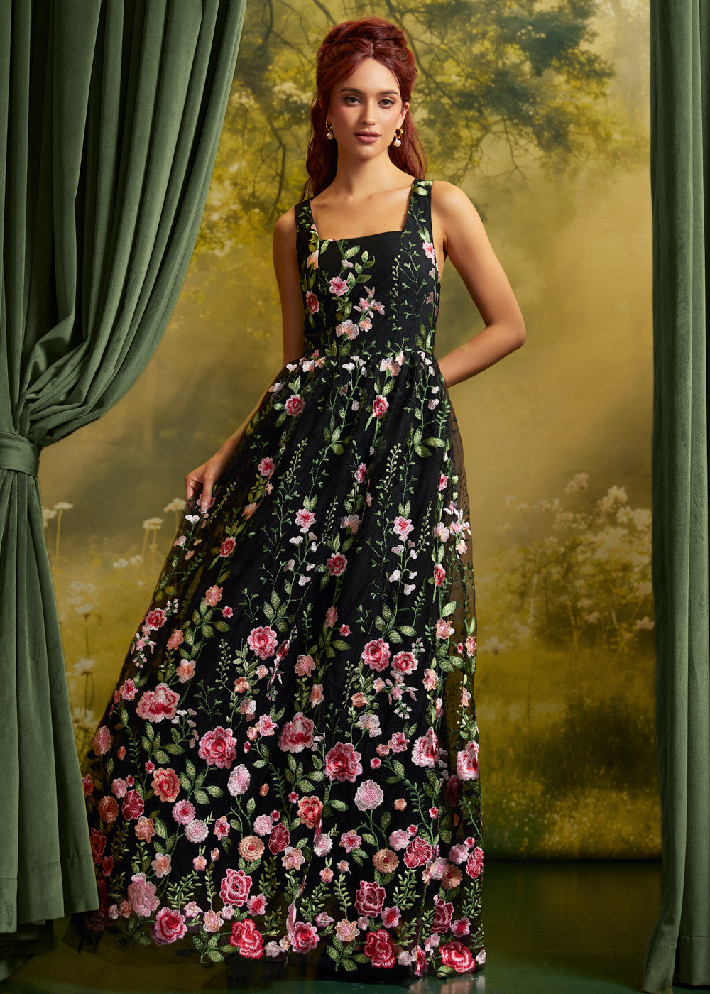 A woman models a sleeveless black maxi dress with a vibrant pink and red floral print, set against a scenic backdrop with green curtains.