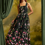 A woman models a sleeveless black maxi dress with a vibrant pink and red floral print, set against a scenic backdrop with green curtains.