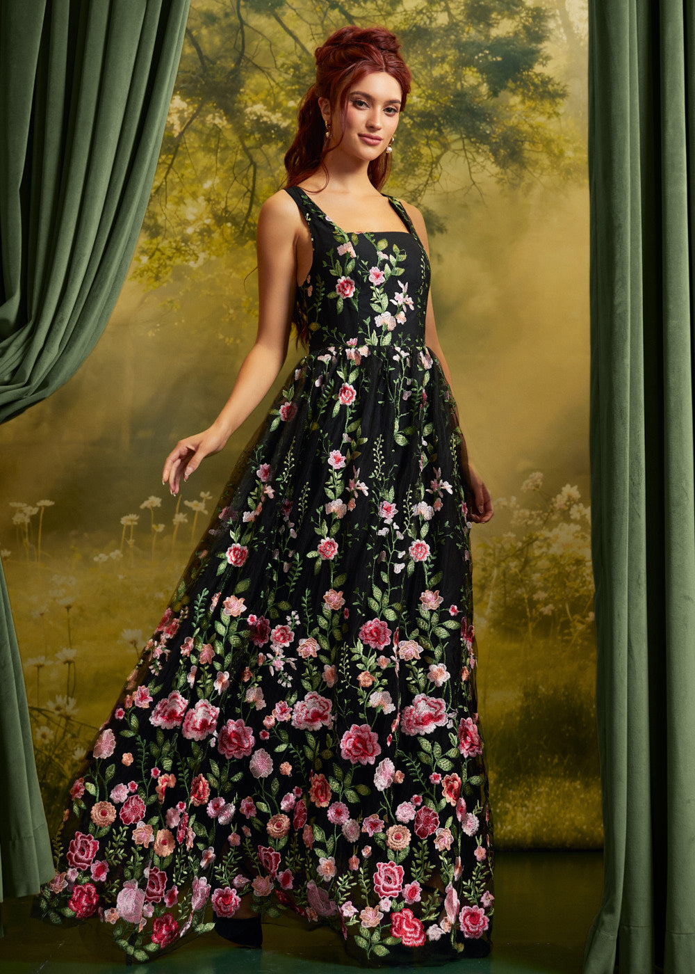 A woman modeling a sleeveless black maxi dress with a vibrant pink and red floral print, set against a moody green and gold forest backdrop with draped curtains.