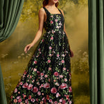 A woman modeling a sleeveless black maxi dress with a vibrant pink and red floral print, set against a moody green and gold forest backdrop with draped curtains.