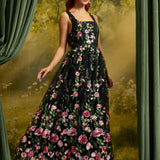 A woman modeling a sleeveless black maxi dress with a vibrant pink and red floral print, set against a moody green and gold forest backdrop with draped curtains.