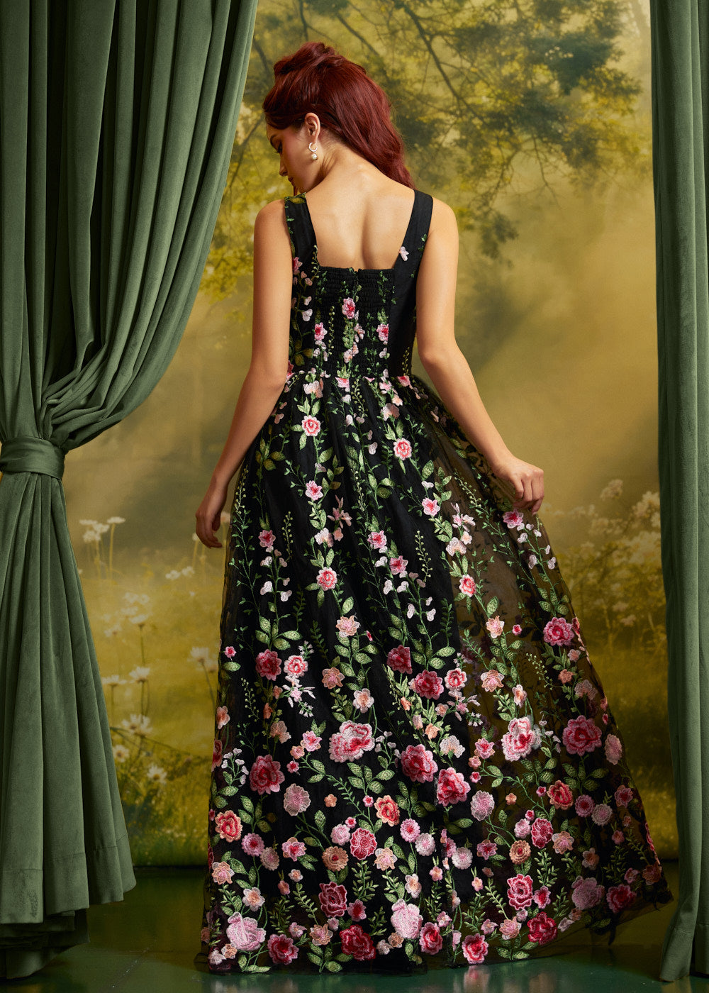 A woman stands with her back to the camera wearing a sleeveless black maxi dress covered in pink, red, and white floral embroidery with green leaves. She is posed in front of a scenic backdrop with green curtains on the sides.