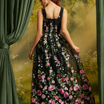 A woman stands with her back to the camera wearing a sleeveless black maxi dress covered in pink, red, and white floral embroidery with green leaves. She is posed in front of a scenic backdrop with green curtains on the sides.