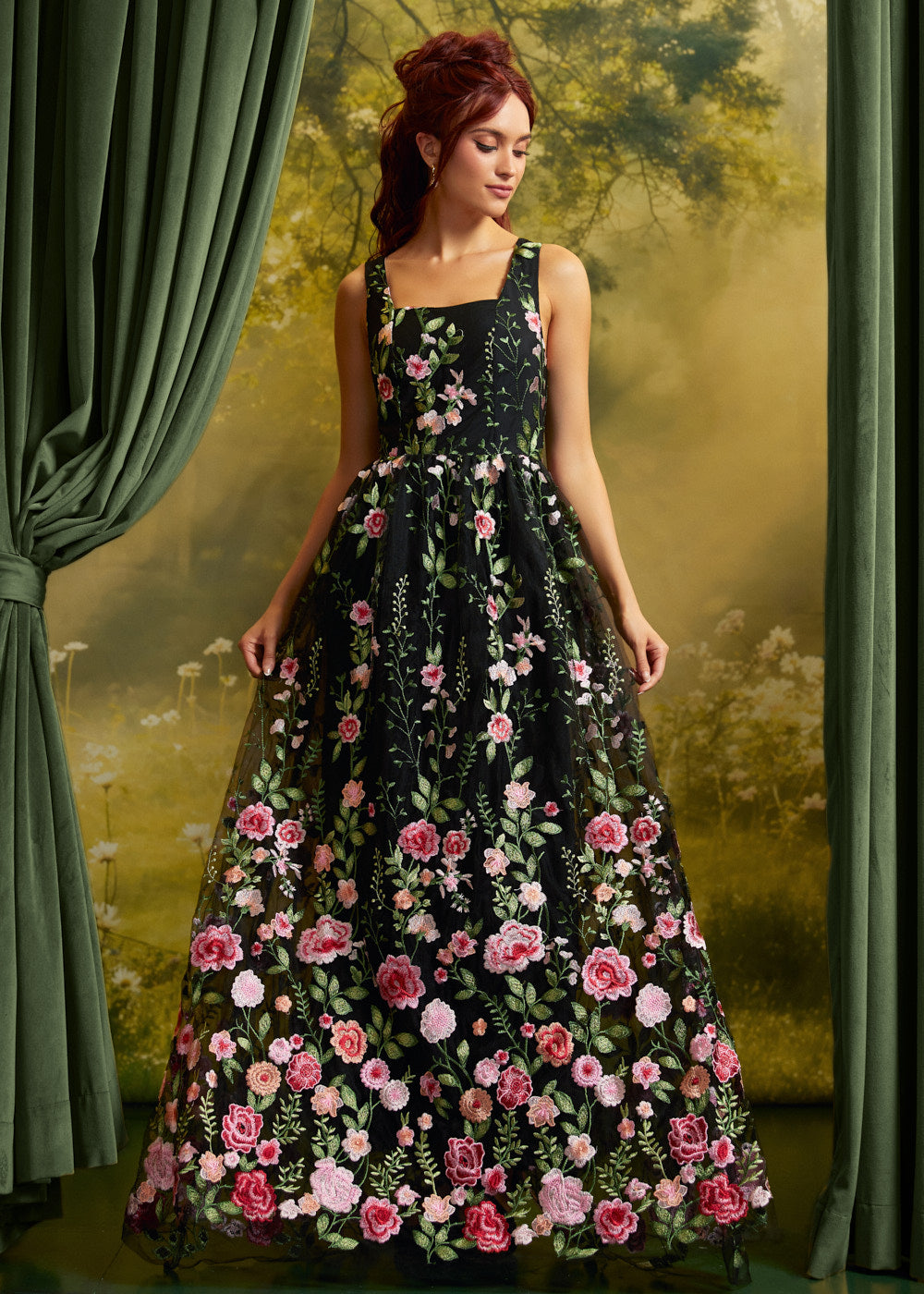 A woman wearing a sleeveless black maxi dress with a vibrant pink and green floral print, posed in front of a vintage-style green curtain backdrop.