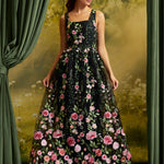 A woman wearing a sleeveless black maxi dress with a vibrant pink and green floral print, posed in front of a vintage-style green curtain backdrop.