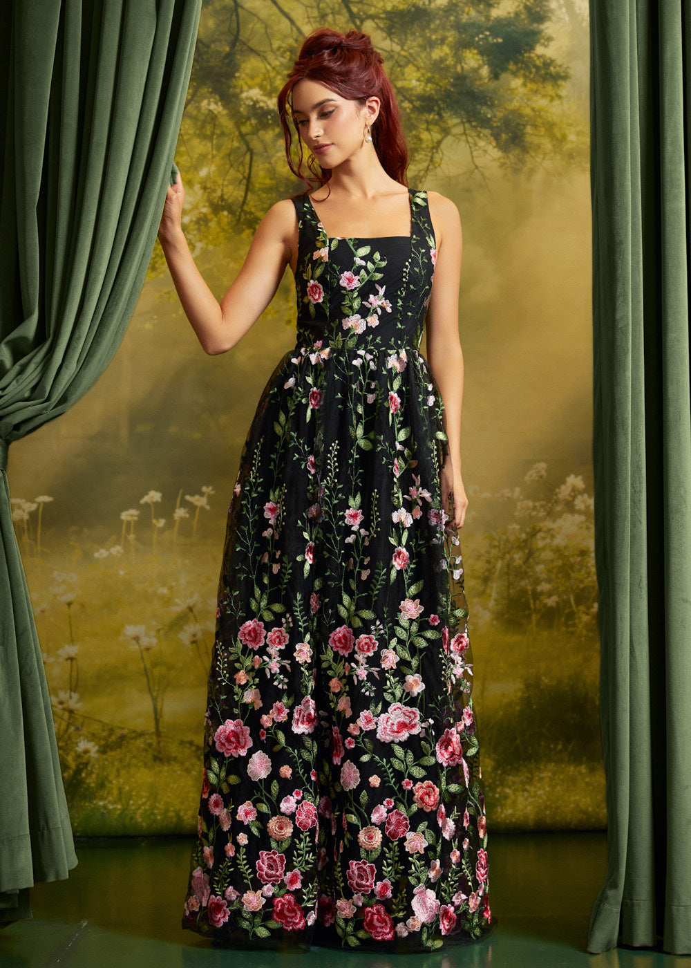 A woman with red hair models a black sleeveless maxi dress with a pink and green floral print, standing in front of a vintage-style forest backdrop with green curtains.
