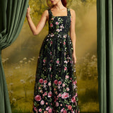 A woman with red hair models a black sleeveless maxi dress with a pink and green floral print, standing in front of a vintage-style forest backdrop with green curtains.