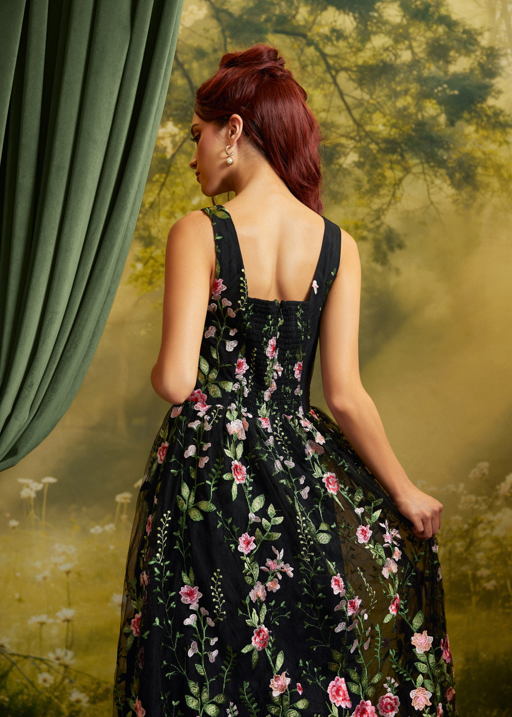 A woman with red hair is shown from the back wearing a sleeveless black ball gown with a pink and green floral pattern. She is posed in front of a vintage-style backdrop featuring a green curtain on the left and a yellow-green forest scene.