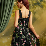 A woman with red hair is shown from the back wearing a sleeveless black ball gown with a pink and green floral pattern. She is posed in front of a vintage-style backdrop featuring a green curtain on the left and a yellow-green forest scene.