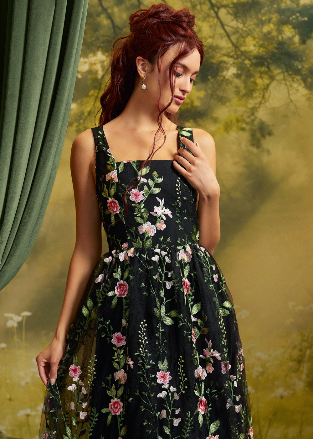 A woman with red hair wearing a sleeveless black midi dress with pink and green floral embroidery, posed against a vintage-style green and yellow backdrop.