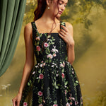 A woman with red hair wearing a sleeveless black midi dress with pink and green floral embroidery, posed against a vintage-style green and yellow backdrop.