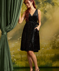 Model wearing a black velvet faux-wrap dress with a tied waist sash and black strappy heels, with one hand tucked into a side pocket beside a green curtain.