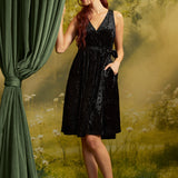Model wearing a black velvet faux-wrap dress with a tied waist sash and black strappy heels, with one hand tucked into a side pocket beside a green curtain.
